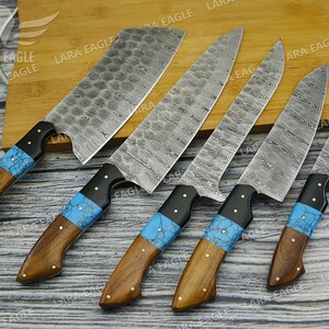 May include: Five kitchen knives with blue and black handles and Damascus steel blades. The knives are arranged on a wooden cutting board.