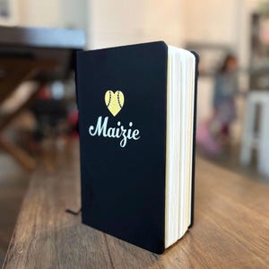 May include: A black notebook with the name "Maizie" in white script, beneath a gold heart-shaped baseball design. The notebook is open, revealing the cream-colored pages. The notebook is on a wooden surface.