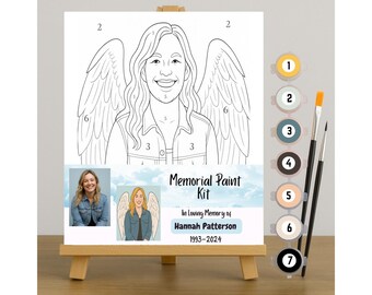 May include: A paint-by-numbers kit on a small wooden easel. The kit features a line drawing of a person with angel wings, with numbers indicating paint colors. The text reads "Memorial Paint Kit" and "In Loving Memory of Hannah Patterson 1993-2024."