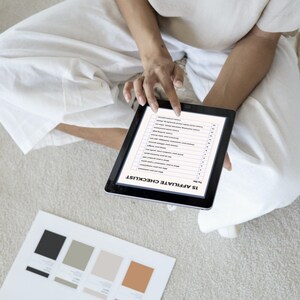 Op de afbeelding: Een persoon zit op een lichtgekleurd tapijt en gebruikt een zwarte tablet met een checklist met de titel "15 Affiliate Checklist". Kleurstalen en witte dozen zijn in de buurt zichtbaar. De persoon draagt witte kleding.