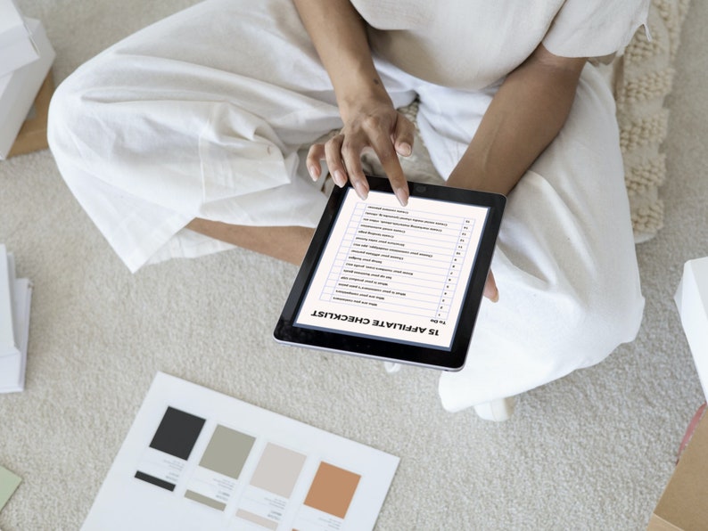 Op de afbeelding: Een persoon zit op een lichtgekleurd tapijt en gebruikt een zwarte tablet met een checklist met de titel "15 Affiliate Checklist". Kleurstalen en witte dozen zijn in de buurt zichtbaar. De persoon draagt witte kleding.
