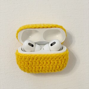 May include: A pair of white wireless earbuds inside an open white charging case, encased in a bright yellow crocheted cover. The case is set against a textured white background, showcasing the handmade, protective accessory.