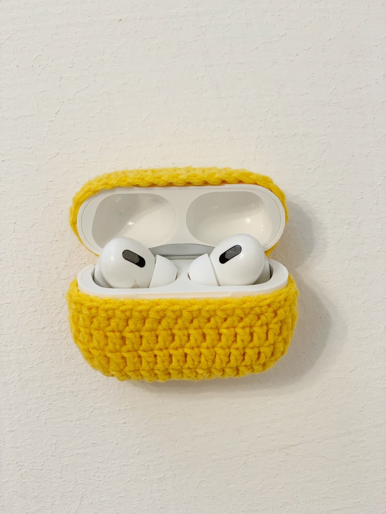 May include: A pair of white wireless earbuds inside an open white charging case, encased in a bright yellow crocheted cover. The case is set against a textured white background, showcasing the handmade, protective accessory.