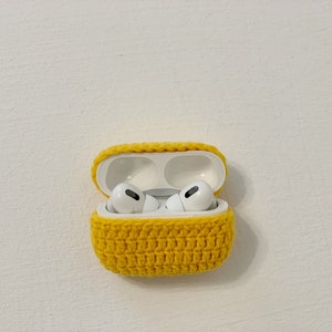 May include: A yellow crocheted case for AirPods Pro, open to reveal the white earbuds inside. The case is made of a textured, hand-stitched yarn and sits against a white background.
