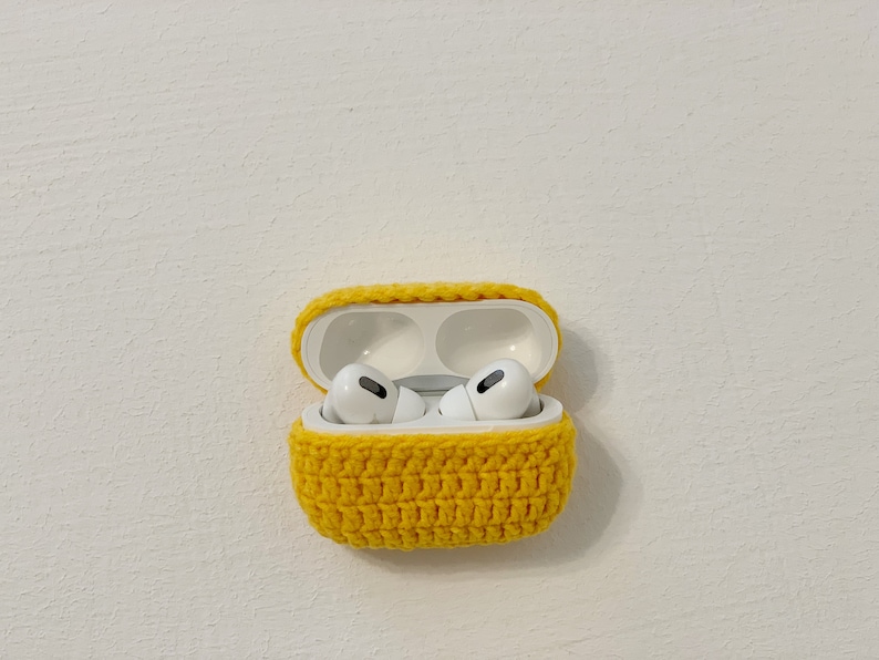 May include: A yellow crocheted case for AirPods Pro, open to reveal the white earbuds inside. The case is made of a textured, hand-stitched yarn and sits against a white background.