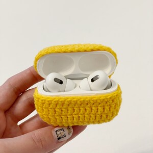 May include: A bright yellow, crocheted case for wireless earbuds. The case is open, revealing white earbuds inside. The case is held in a hand against a white background. The crochet work is visible, with a textured appearance.