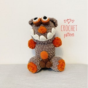 Puede incluir: Un juguete de monstruo de ganchillo hecho a mano en tonos grises, naranjas y marrones. La criatura de peluche tiene grandes dientes blancos, ojos naranjas y detalles naranjas en los pies y los brazos. El texto "CROCHET pattern" está escrito en rojo.