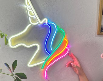 Può includere: Una testa di unicorno in neon bianco con una criniera arcobaleno. L'unicorno è montato su una parete bianca.