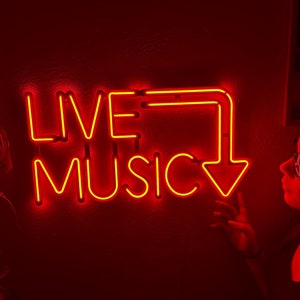 Può includere: Un cartello al neon rosso con le parole "LIVE MUSIC" in un font al neon rosso. Il cartello è su una parete rosso scuro. Una persona è in piedi a destra del cartello, guardandolo.