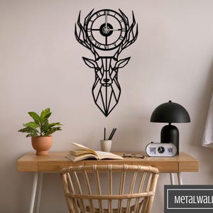 May include: Black metal geometric deer head wall clock with a clock face in the center of the antlers. The clock is mounted on a white wall above a wooden desk with a lamp, a plant, and a book.