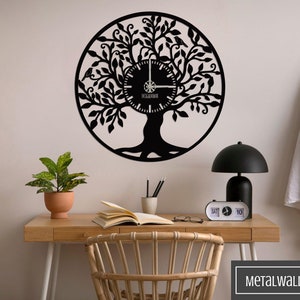 May include: Black metal wall clock in the shape of a tree of life with intricate details. The clock is mounted on a white wall above a wooden desk with a potted plant, books, and glasses. The clock has a black background and white hands and numbers.