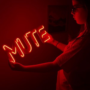 May include: A glowing red neon sign spelling out the word "MUTE" is held in front of a dark red background. The neon sign is illuminated, casting a warm glow. The sign is held by a person.
