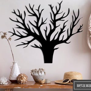 Könnte beinhalten: Schwarze Wandkunst aus Metall in Form eines Baumes mit kahlen Ästen. Die Kunst ist an einer weißen Wand in einem Raum mit einer Holzleiter, einem Korbstuhl und Dekorationsgegenständen auf einem Holztisch angebracht.