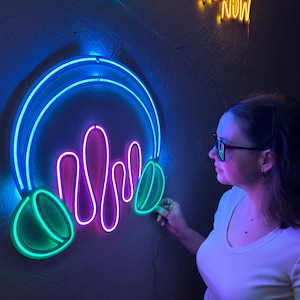 May include: A neon sign in the shape of headphones with a sound wave design. The sign features a blue outline, with green and purple accents. The sign is mounted on a dark wall.