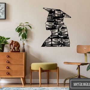 May include: Black metal wall art depicting a person wearing a graduation cap, made of stacked books. The art is mounted on a beige wall with a clock, a wooden dresser, a green plant, and a wooden stool in the background.