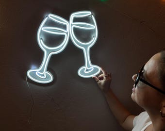 May include: A white neon sign depicting two wine glasses clinking together. The sign is mounted on a wall and is lit up.