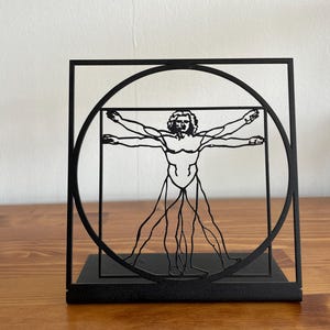 May include: Black metal sculpture of a human figure in a square frame with a circle around it. The figure is in the Vitruvian Man pose with arms and legs outstretched.