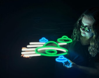 May include: A neon sign shaped like a flying saucer with green and blue lights. The sign is held by a person in a dark room.