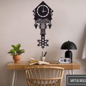 May include: Black metal wall clock with intricate details, featuring a clock face with Roman numerals and a pendulum. The clock is mounted on a white wall above a wooden desk with a lamp, a plant, and a book.