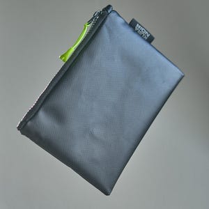 Könnte beinhalten: Eine rechteckige schwarze Reißverschlusstasche mit strukturierter Oberfläche. Die Tasche verfügt über einen leuchtend lindgrünen Reißverschlussanhänger und ein kleines schwarzes Stoffetikett an der Seite mit dem weißen Text KURA NOIR.