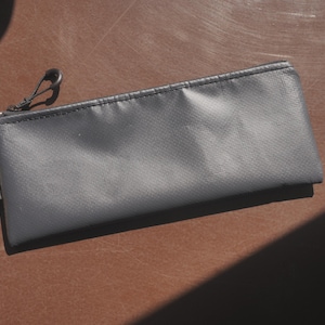 Può includere: Un astuccio grigio con chiusura a cerniera e una piccola etichetta nera con la scritta "Kura Noir". L'astuccio rettangolare è realizzato in un materiale strutturato e si trova su una superficie marrone.