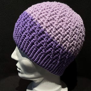 Könnte beinhalten: Eine gestrickte Mütze in Lila und Lavendel mit einem strukturierten Stichmuster. Die Mütze wird auf einem weißen Mannequin-Kopf getragen.