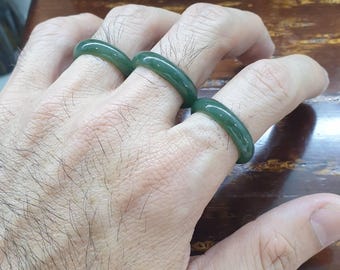 May include: Three dark green jade rings are worn on the fingers of a hand. The rings have a smooth, polished surface and a rounded shape. The background includes a wooden surface and two statues.
