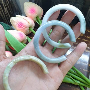 May include: Three jade bangles in shades of green and blue are displayed. The smooth, curved bracelets are held in a hand, with pink and white tulips and green stems in the background. The bangles are a classic jewelry item.