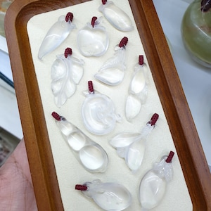 Könnte beinhalten: Eine Sammlung von zehn weißen Jade-Anhängern in Form von Obst und Gemüse, die auf einem Holztablett mit beigefarbenem Filz ausgelegt sind. Die Anhänger haben rote Schnur-Schlaufen zum Aufhängen.