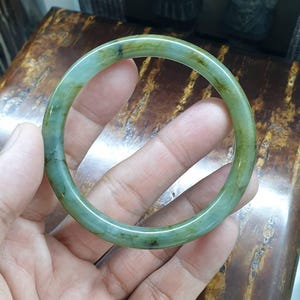 May include: A jade bangle bracelet in shades of green and brown is held in a hand. The smooth, polished surface of the bracelet reflects light, highlighting its natural variations. The bracelet's circular shape is clearly visible.