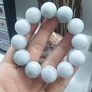 May include: A bracelet featuring large, round, pale green jade beads. The beads are strung together, forming a circular shape. The bracelet is held in a hand, showcasing the smooth, polished surface of the jade.
