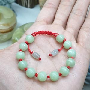 May include: A light green beaded bracelet with a red cord. The bracelet features round, polished beads and a sliding knot closure. The beads are approximately 0.5 inches in diameter.