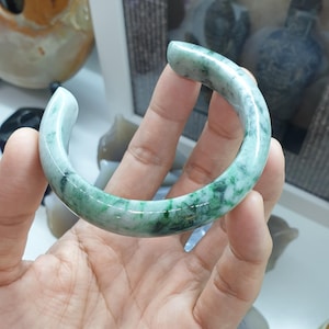 May include: A jade bangle bracelet with a marbled green and white pattern. The smooth, polished surface of the bracelet is curved, with rounded ends. The bracelet is held in a hand.