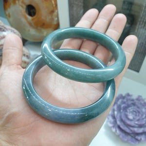 May include: Two jade green bangle bracelets, one slightly larger than the other, are stacked on top of each other. The bracelets are smooth and polished.