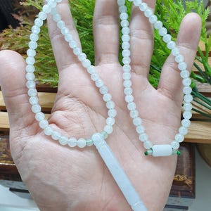 May include: A necklace featuring a strand of small, pale green, translucent beads. The necklace includes a long, cylindrical, pale green pendant and a small, rectangular, pale green bead connector, both secured with thin green cord accents.