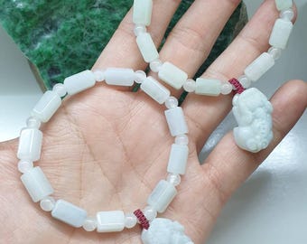 Może przedstawiać: Dwie bransoletki z bladozielonego jadeitu z prostokątnymi koralikami i małymi okrągłymi przekładkami. Każda bransoletka zawiera rzeźbioną zawieszkę z jadeitu w kształcie mitycznego stworzenia, przymocowaną ciemnoczerwonym sznurkiem, tworząc zestaw.