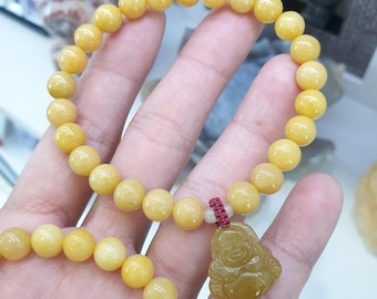 Op de afbeelding: Een gele jade armband met een gele jade hanger van een lachende Boeddha. De armband is gemaakt van ronde kralen en heeft een rode touwtjesluiting.