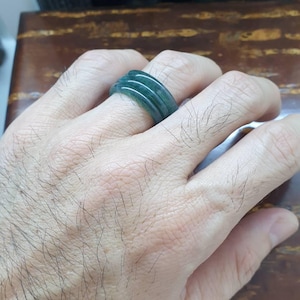 Puede incluir: Un anillo de jade verde oscuro con tres bandas apiladas. El anillo es liso y pulido, reflejando la luz. El anillo es una banda sólida, sin roturas ni engastes visibles. El anillo es una joya sencilla y elegante.
