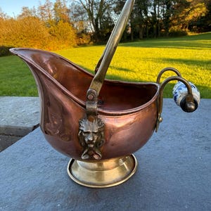 May include: A copper coal scuttle featuring a brass pedestal base and a brass handle attached with lion head mounts. The rear handle includes a white and blue floral patterned ceramic grip. The vessel has a wide, open top and a curved spout.