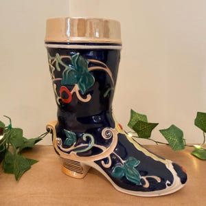 May include: A ceramic boot-shaped vessel featuring a glossy dark blue glaze with raised relief details of green ivy leaves, swirling cream-coloured vines, and small red accents. The top rim and the block heel have a metallic gold-toned finish.