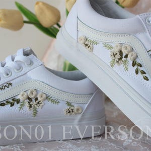 Könnte beinhalten: Ein Paar weiße Canvas-Sneaker mit gestickten Blumen und grünen Blättern. Ein Schuh hat den Text "Frau [Nachname]" an der Seite gestickt. Der andere Schuh hat den Text "D4-10-24" an der Seite gestickt.