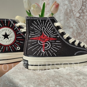 Może przedstawiać: Czarne, wysokie buty Converse z białymi szwami i czerwonym haftem przedstawiającym węża i krzyż. Buty mają białą gumową podeszwę, a logo Converse All Star jest wyszyte z boku.
