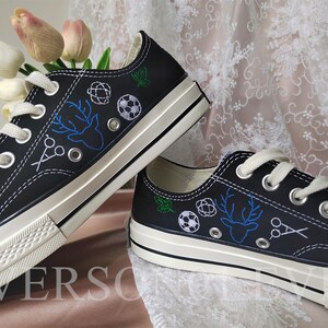 May include: A pair of black canvas shoes with white stitching and embroidered designs. The designs include a deer, scissors, a soccer ball, and a circle with a star inside.