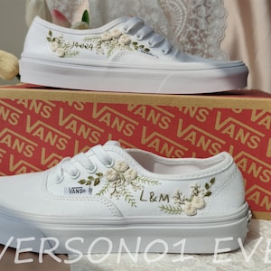 Könnte beinhalten: Weiße Canvas-Sneaker mit weißen Schnürsenkeln und gestickten Blumen und Grün. Die Sneaker haben den Text "L&M" an der Seite gestickt. Die Sneaker stehen auf einem Karton mit dem Wort "VANS" darauf.