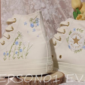 Puede incluir: Zapatillas altas color crema con bordados florales. Las zapatillas presentan flores bordadas azules y blancas y ojales dorados. El logotipo de Converse All Star es visible en el lateral. Los zapatos se exhiben sobre una superficie de madera.