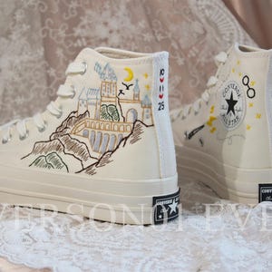May include: Cream-colored high-top sneakers with custom embroidery. One shoe features a castle design with a moon and birds, while the other has a star and symbols. The shoes have white laces and black soles.