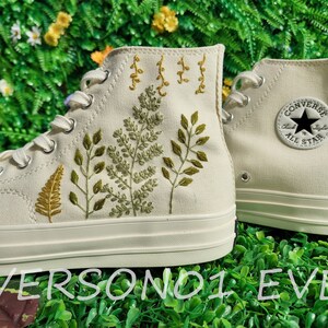 May include: A pair of white high-top sneakers with green and yellow embroidered leaves and the Converse logo on the side.