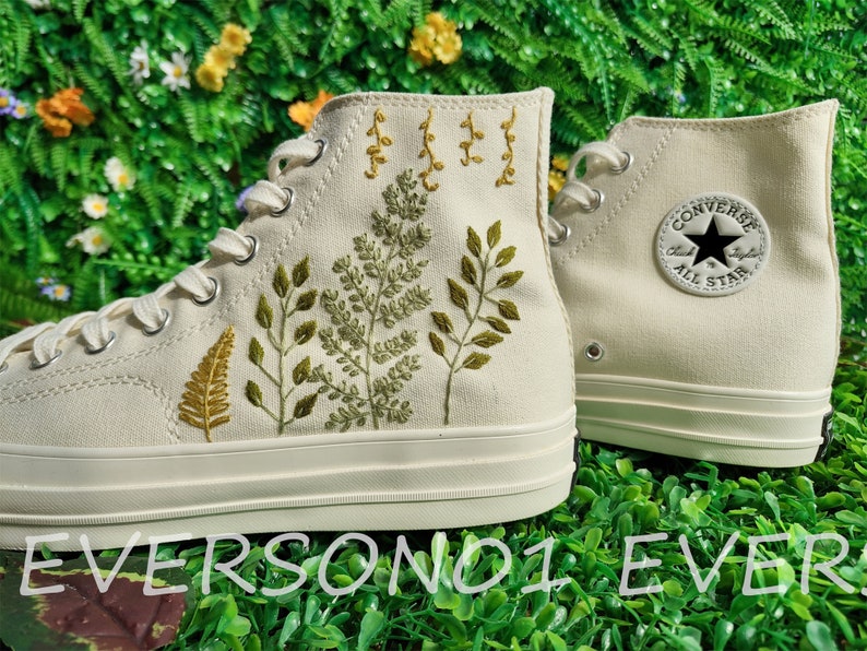 May include: A pair of white high-top sneakers with green and yellow embroidered leaves and the Converse logo on the side.