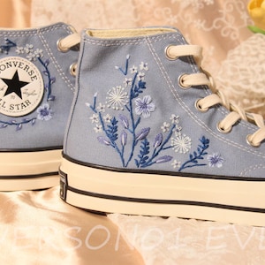 Puede incluir: Un par de zapatillas de lona altas de color azul claro con cordones blancos y suela de goma blanca. Las zapatillas están bordadas con un diseño floral en tonos azules y blancos. La zapatilla izquierda tiene un parche con el logotipo de Converse All Star.