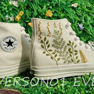 May include: White high-top Converse sneakers with green and yellow embroidered floral designs. The sneakers have a black Converse logo and the text "Chuck Taylor All Star" on the side.